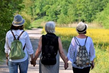 Three women are seen from behind, walking hand in hand along a path in an outdoor setting